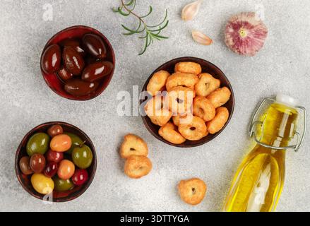 Tarallini-petits bagels italiens traditionnels. Taralli. Collation savoureuse. Olives noires et vertes, Kalamata, huile d'olive, ail et romarin sur béton gris Banque D'Images