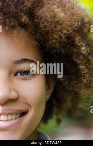 Visage aligné à gauche avec les cheveux bouclés pleins souriant et montrant des dents avec des boucles d'oreilles cloutées dans le jardin Banque D'Images