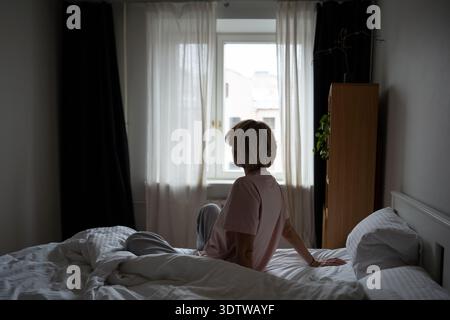 Femme calme assise au lit tôt le matin après le réveil, regardant la fenêtre profitant de la lumière du jour. Banque D'Images