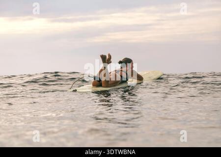 Jeune femme reposant sur un surf dans la mer calme, jambes levées et détendues, Scanning Soft Clouded Horizon, patient Banque D'Images