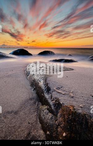 Une photographie longue exposition à couper le souffle capturant un coucher de soleil serein sur une plage rocheuse. Le ciel est orné de nuages dramatiques et striés peints de teintes Banque D'Images