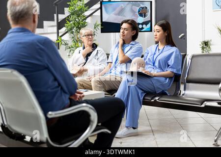 Le jeune homme adulte explique les symptômes aux médecins, exprimant la tension musculaire et l'inconfort lors de la consultation hospitalière de routine. Le patient masculin est assis dans la salle d'attente de la clinique avec le personnel médical féminin. Banque D'Images