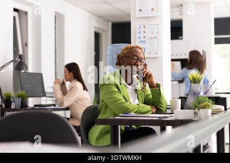 Femme noire s'engage dans l'appel téléphonique du client au bureau, finalisant l'accord commercial réussi. Une professionnelle féminine souriante réussit le projet grâce à une communication efficace et à un professionnalisme. Banque D'Images