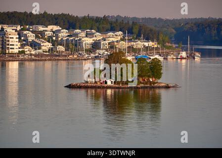 Une scène idyllique mettant en vedette une petite île rocheuse couverte d'arbres avec quelques pittoresques bâtiments rouges et blancs au premier plan, contre le calme, refl Banque D'Images