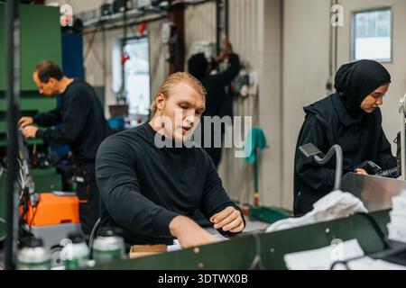 Jeune homme col bleu assis à table tout en travaillant en usine Banque D'Images
