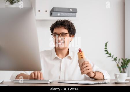 Jeune homme d'affaires avec hot dog savoureux travaillant dans le bureau Banque D'Images