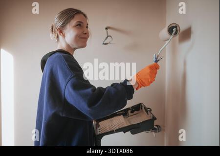 Une jeune femme peint en toute confiance le mur d'une pièce à l'aide d'une brosse rotative. Portant des gants de protection et tenant un bac à peinture, elle ajoute un nouveau manteau à Banque D'Images