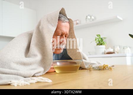 Homme mûr avec serviette faisant inhalation de vapeur à table dans la cuisine Banque D'Images