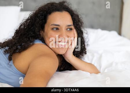 Femme afro-américaine adulte couchée dans le lit, reposant la tête sur la main portant un débardeur bleu clair Banque D'Images