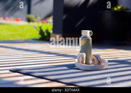 La bouteille d'eau texturée et les écouteurs beiges reposent sur un tapis sombre sur la terrasse ensoleillée, avec des bandes moulées Banque D'Images