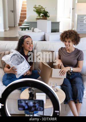 Femmes afro-américaines déballant l'appareil enveloppé de bulles sur le canapé filmant avec la lumière annulaire et le téléphone Banque D'Images