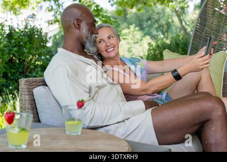 Couple diversifié assis sur un canapé en osier dans le jardin, femme tenant un smartphone prenant selfie avec des boissons Banque D'Images