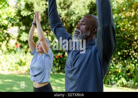 Couple diversifié pratiquant le yoga debout étiré sur la pelouse dans le parc portant montre de fitness et leggings Banque D'Images