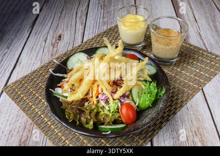 Une salade japonaise composée de légumes frais recouverts de tempura de petits poissons croustillants, servie avec deux sortes de vinaigrettes sur une table rustique en bois. Banque D'Images