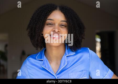 Femme afro-américaine souriante, debout dans l'entrée portant un haut en satin bleu, cerceaux en or, porte Banque D'Images