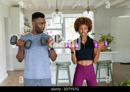 Couple afro-américain curling haltères dans la cuisine de la maison portant un tee-shirt bleu et des leggings magenta Banque D'Images
