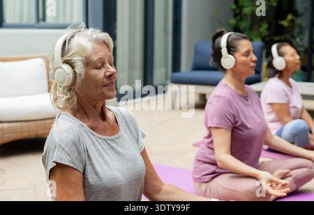 Diverses femmes âgées et matures méditant sur le patio portant des écouteurs blancs, des tapis de yoga, un espace de copie Banque D'Images