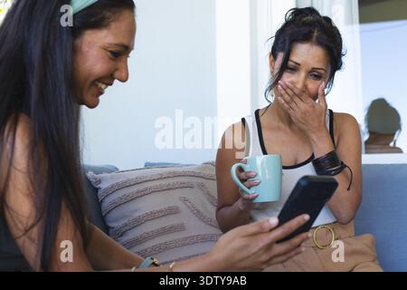 Diverses amies féminines assis sur le canapé à la maison, femme gauche tenant le téléphone, femme droite sirotant la tasse Banque D'Images