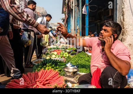 Un homme est assis à un marché de rue dans le vieux Delhi, vendant divers légumes frais. D'autres acheteurs et stands de marché sont visibles au loin. Banque D'Images