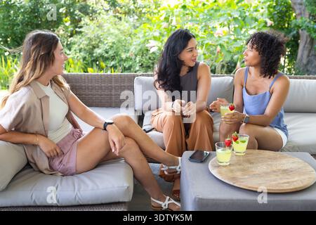 Diverses amies féminines discutant sur le patio portant des tenues avec des sandales, des cocktails et un smartphone Banque D'Images