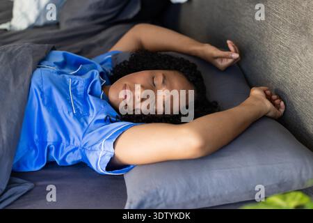 Femme afro-américaine allongée sur le lit, portant un dessus bleu clair, reposant sur une literie grise, plante visible Banque D'Images