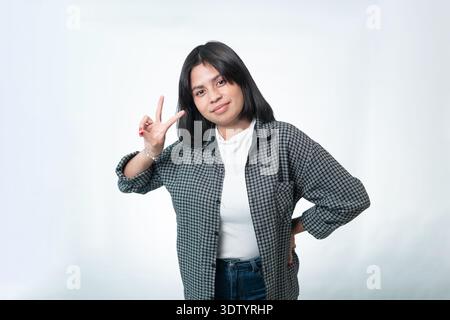 Portrait d'une femme indonésienne heureuse donnant le signe V ou le geste de paix. Photographie de style de vie décontracté avec une chemise à carreaux sur un fond blanc Banque D'Images