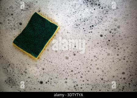 Éponge de cuisine verte et jaune flottant dans une mousse de savon épaisse avec des bulles visibles. Vue rapprochée mettant en évidence la texture, l'humidité et l'action de nettoyage. Banque D'Images