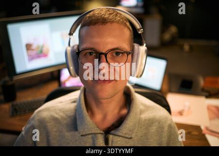 Portrait de jeune homme caucasien adulte portant des lunettes et des écouteurs regardant dans la caméra, assis au bureau avec des moniteurs d'ordinateur affichant des œuvres d'art numériques dans l'espace de travail créatif Banque D'Images