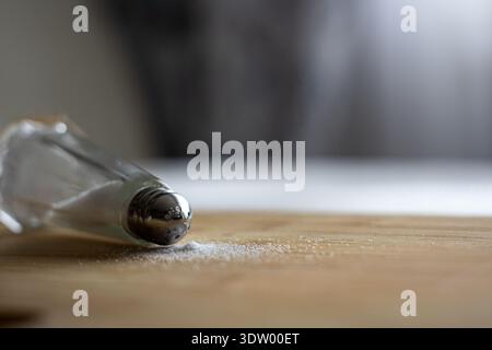 Un gros plan artistique d'une salière en verre classique couchée sur le côté sur une planche à découper en bambou ou en bois. Une petite quantité de sel blanc contient spil Banque D'Images