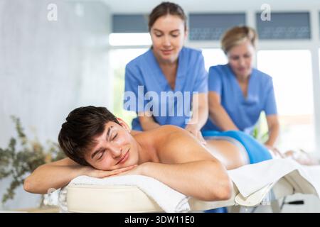 Jeune homme relaxant pendant que deux masseuses font un massage du dos à quatre mains Banque D'Images