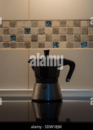 Gros plan de la cafetière Moka pot sur le poêle à induction noir contre le mur de carreaux de mosaïque gris en basse lumière Banque D'Images