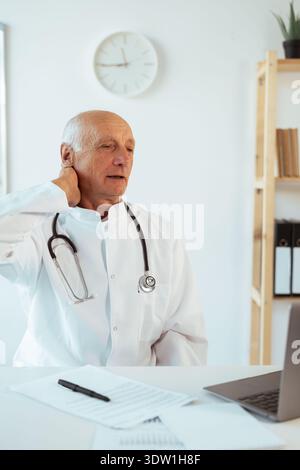 Médecin homme senior travaillant sur ordinateur portable et souffrant de douleurs au cou, stressé ou épuisé sur le lieu de travail à l'hôpital. Professionnel médical masculin souffrant de Banque D'Images