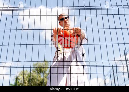 Femme urbaine avec basket-ball regardant à travers la clôture grise sur le terrain de basket-ball extérieur. Banque D'Images