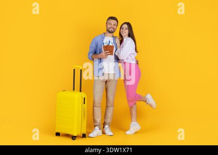 Couple souriant avec passeports debout par valise jaune sur fond de studio lumineux Banque D'Images
