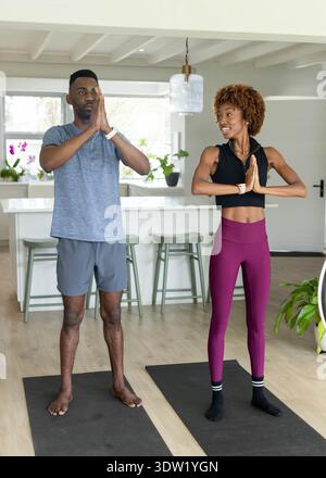 Couple diversifié pratiquant le yoga debout sur des tapis dans la cuisine, portant des vêtements de sport et des montres Banque D'Images