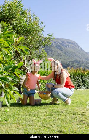 Femme et fille enfant portant des oreilles de lapin collectant des oeufs colorés dans un panier en osier sur la pelouse Banque D'Images