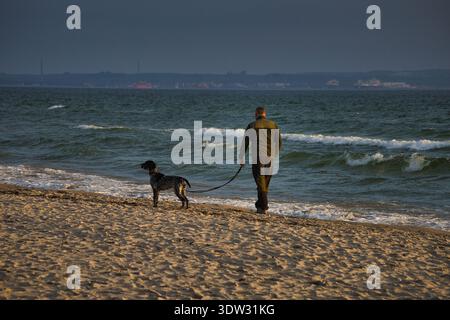 Promenade matinale sur la plage de Binz : un homme méconnaissable promène son chien en laisse le long des vagues de la mer Baltique sur l'île de Ruegen, en Allemagne Banque D'Images