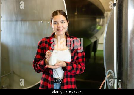 Jeune femme positive travaillant dans une ferme laitière debout avec un verre de lait Banque D'Images