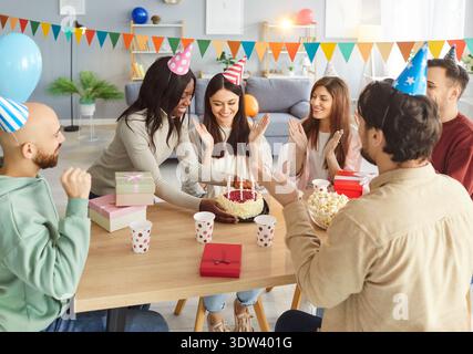 Groupe de jeunes heureux portant des chapeaux de fête applaudissant et félicitant la femme d'anniversaire avec un gâteau. Banque D'Images