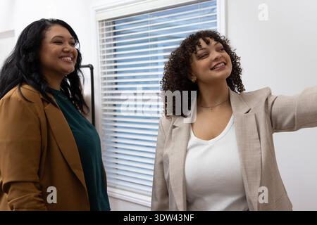 Diverses collègues féminines souriantes et debout ensemble dans le bureau avec blazers, stores, rack Banque D'Images
