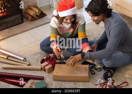 Couple diversifié assis sur le tapis près de la cheminée enveloppant des cadeaux avec chapeau de Père Noël et des rubans Banque D'Images