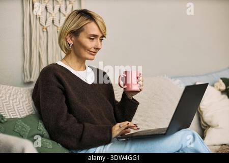 Femme mature caucasienne assise sur un canapé à l'aide d'un ordinateur portable et d'une tasse de maintien, s'engageant avec la plate-forme de santé femtech, en se concentrant sur les solutions de bien-être numériques dans l'environnement domestique Banque D'Images