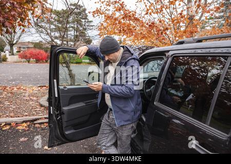 Homme debout près de la porte de voiture ouverte et vérifiant le smartphone à l'extérieur en automne Banque D'Images