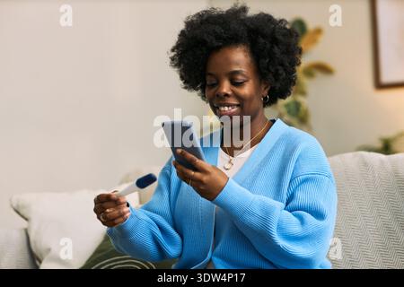 Jeune adulte femme noire assise sur un canapé souriant tout en tenant un smartphone dans une main et un test d'ovulation numérique dans l'autre main, en utilisant la technologie de santé femtech à la maison Banque D'Images