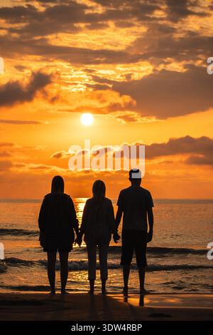 Trois personnes se tenant la main et regardant le lever du soleil sur la mer, debout pieds nus sur la plage sous les nuages dorés du matin. Banque D'Images