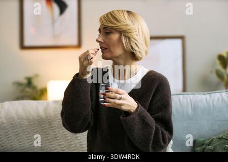 Femme caucasienne d'âge moyen assise à l'intérieur tenant un verre d'eau et prenant la pilule, démontrant l'auto-soin et la gestion de la santé, expression ciblée Banque D'Images