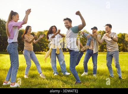Heureux amis dansant acclamant avec enthousiasme dans le parc verdoyant ensoleillé, groupe diversifié applaudissant et célébrant Banque D'Images