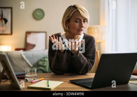 Femme caucasienne d'âge moyen assise au bureau faisant des gestes tout en ayant une consultation en ligne à l'aide d'un ordinateur portable dans le cadre de la maison, discutant des sujets de santé fémintech avec bloc-notes visible et verre d'eau Banque D'Images