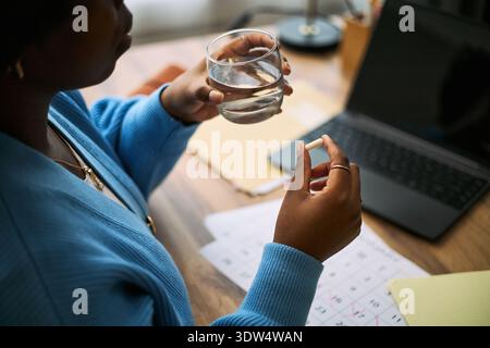 Jeune femme noire adulte tenant la pilule et le verre d'eau près du calendrier et de l'ordinateur portable, se préparant à prendre des médicaments, illustrant la gestion de la santé de la femme et le suivi quotidien du bien-être Banque D'Images