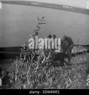 Photographie historique en noir et blanc des années 1970 capture une perspective unique de la ferme piscicole de Donrybkombinat dans le district de Slavyansk, région de Donetsk, RSS d'Ukraine, URSS. La composition présente une végétation verdoyante au bord de l'étang au premier plan, avec des figures floues de pêcheurs dévoués travaillant dans un grand bateau en bois en arrière-plan. Cette image d'archives authentique reflète magnifiquement le travail quotidien, le paysage naturel et l'atmosphère calme dans le Donbass paisible avant la guerre, mettant en évidence l'industrie agricole régionale. Banque D'Images
